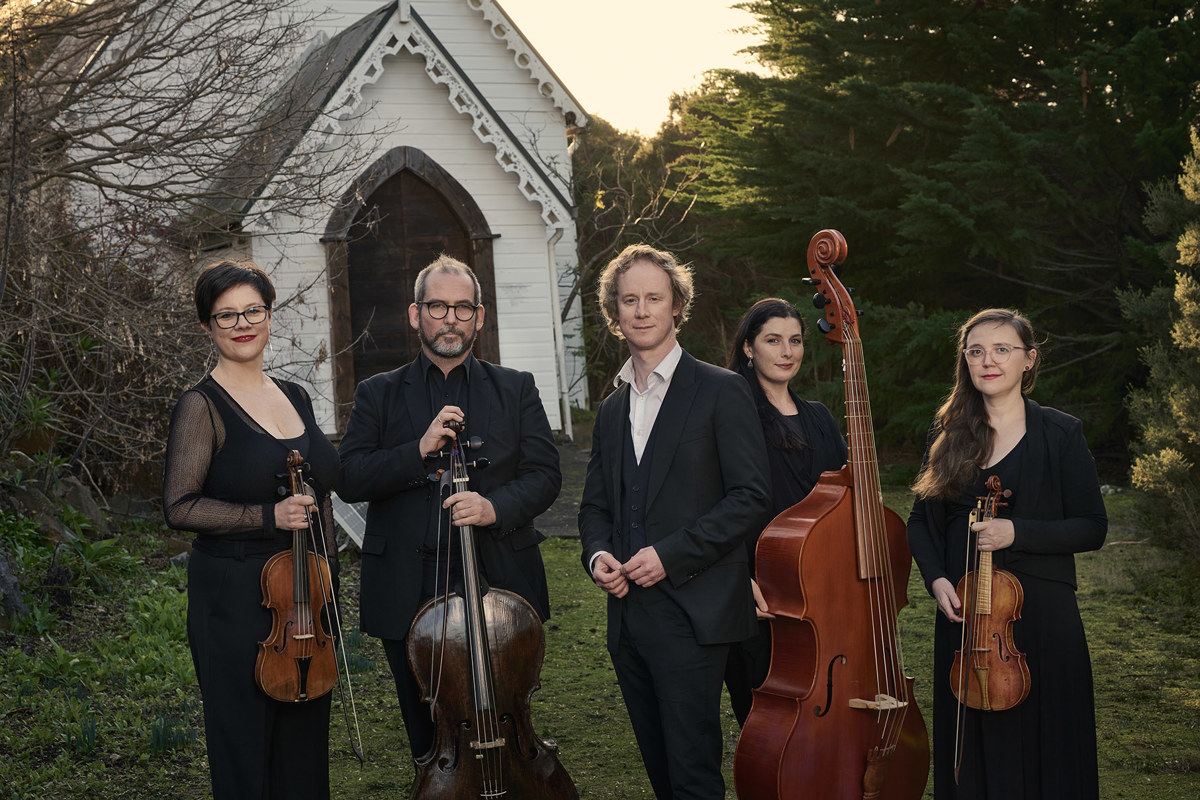A group of five people hold musical instruments standing on the grass in front of an old white small church, this church is surrounded by bushland