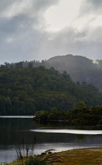 there is a glistening body of water, under beautiful sunlight, a wild mountainous backdrop and a yellow grass spot of land in the forground