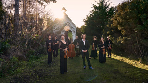 A group of people hold musical instruments standing on the grass in front of an old white small church, this church is surrounded by bushland