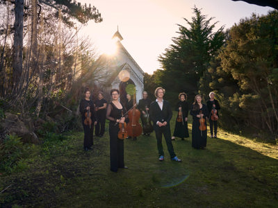 A group of people hold musical instruments standing on the grass in front of an old white small church, this church is surrounded by bushland
