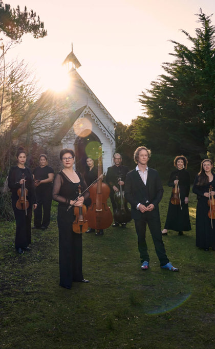 A group of people hold musical instruments standing on the grass in front of an old white small church, this church is surrounded by bushland