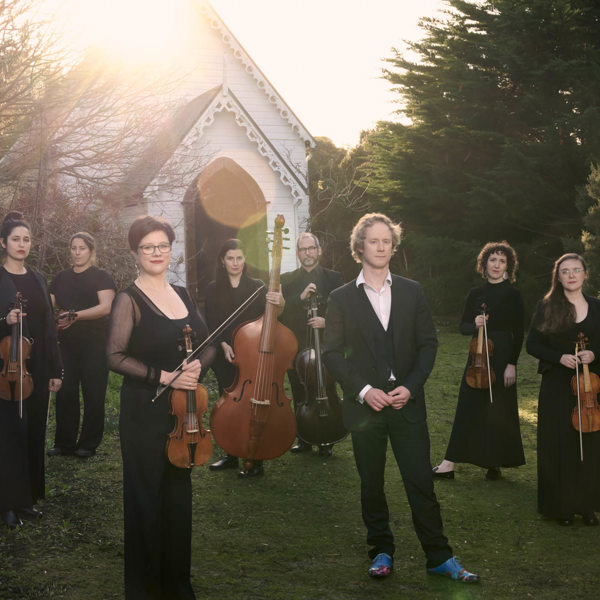 A group of people hold musical instruments standing on the grass in front of an old white small church, this church is surrounded by bushland