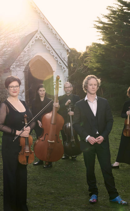 A group of people hold musical instruments standing on the grass in front of an old white small church, this church is surrounded by bushland